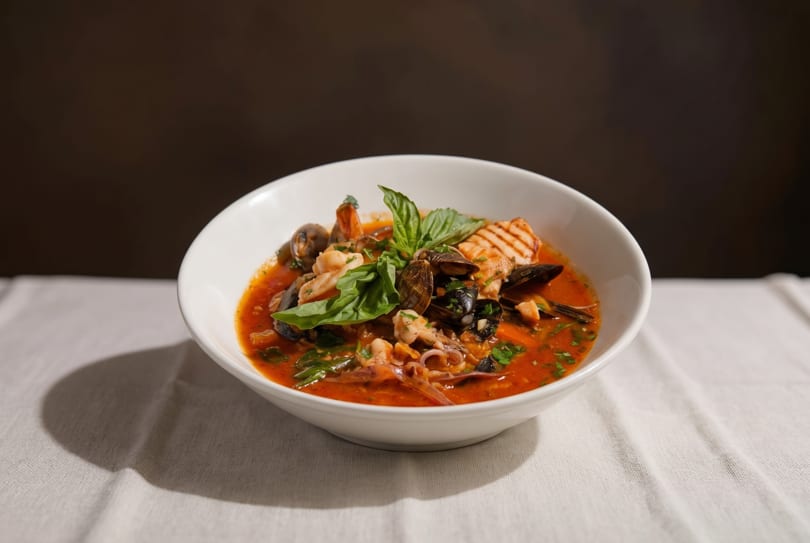 A white bowl of seafood cioppino with mussels, shrimp, fish, and basil on a white tablecloth.