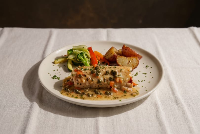Fish with caper sauce, roasted potatoes, and mixed vegetables on a white plate.