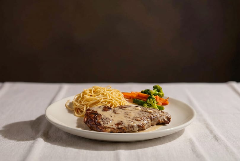 Grilled steak with creamy sauce, spaghetti, and steamed vegetables on a white plate.