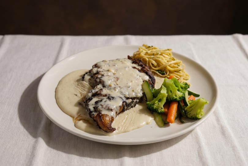 Grilled chicken with creamy sauce, pasta, and steamed vegetables on a white plate.