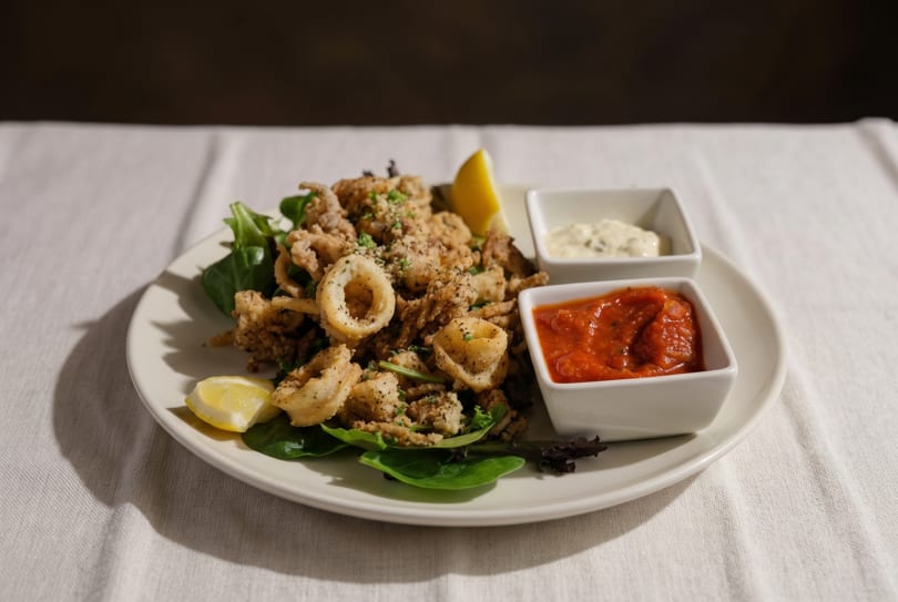 Crispy fried calamari with lemon wedges, marinara sauce, and aioli on a white plate.